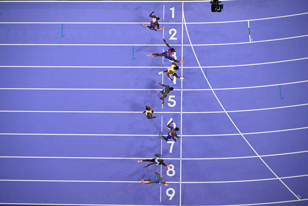 Overhead view of sprinters crossing the finish line in a 100-meter race on a blue track, with the athlete in lane 7 narrowly leading by a small margin.